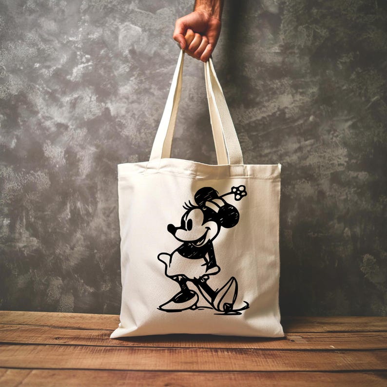 Pode incluir: Saco tote creme com uma ilustra&ccedil;&atilde;o a preto e branco da Minnie Mouse. O saco &eacute; segurado por uma m&atilde;o e tem al&ccedil;as compridas. O gr&aacute;fico da Minnie Mouse &eacute; um design cl&aacute;ssico de desenho animado.