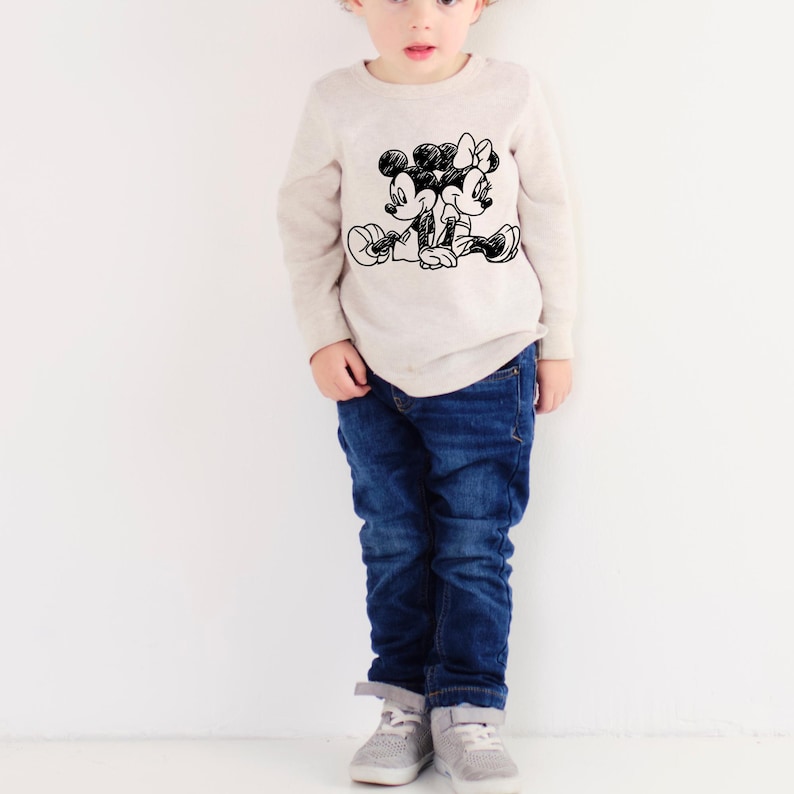 Pode incluir: Uma camisa bege de manga comprida com um esbo&ccedil;o preto de Mickey e Minnie Mouse. A camisa &eacute; combinada com jeans azuis e t&ecirc;nis cinzentos. O fundo &eacute; branco.