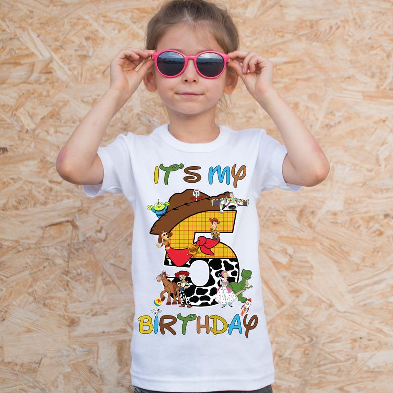 May include: White t-shirt with the text "It's my 5th Birthday" and Toy Story characters. The number 5 is decorated with a cow print and other characters. The shirt is worn by a young girl wearing pink sunglasses.