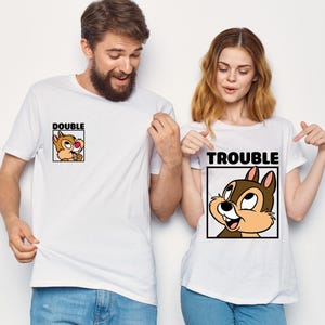 May include: Two white t-shirts with cartoon chipmunk graphics. One shirt says "DOUBLE" with a chipmunk image, the other says "TROUBLE" with a matching chipmunk. The shirts are worn by two people.