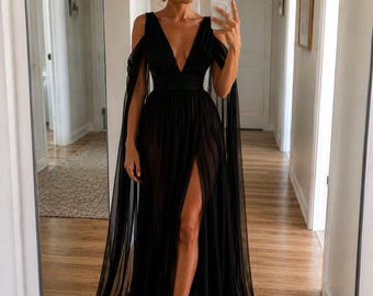May include: A black formal gown with a plunging neckline and a high slit. The dress features flowing cape sleeves and a sheer skirt. The model is wearing silver sandals. The dress is suitable for special occasions.