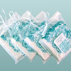 May include: A stack of teal and white cocktail napkins with a floral design are packaged in clear cellophane bags tied with a white ribbon. The label reads "Cocktail Napkins, Hand-Crafted Napkins 5" x 5", The Blue Daisy, &copy; 2014".