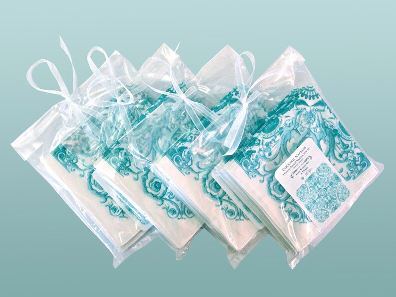 May include: A stack of teal and white cocktail napkins with a floral design are packaged in clear cellophane bags tied with a white ribbon. The label reads "Cocktail Napkins, Hand-Crafted Napkins 5" x 5", The Blue Daisy, &copy; 2014".