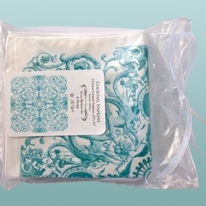 May include: A set of four 10-inch by 10-inch cotton cocktail napkins with a teal and white floral design. The napkins are packaged in a clear plastic bag with a white ribbon. The packaging label reads "Cocktail Napkins, 4 Cotton Cocktail Napkins, 10"x 10", Demon Angela, From Demon Art & Design."