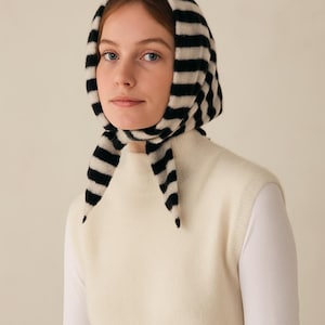 May include: A woman wearing a black and white striped headscarf and a cream-colored sweater. The headscarf is tied under the chin. The sweater is sleeveless and has a high neckline. The background is a solid, neutral color.
