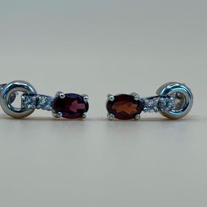 May include: A pair of silver-toned stud earrings. Each earring features a dark red oval gemstone, accented with small clear stones and a circular design. The earrings are displayed against a neutral background.