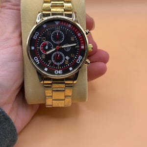 May include: Gold-toned wristwatch with a black dial and red accents. The watch features multiple sub-dials and a gold-toned link bracelet. The watch is displayed on a beige surface.