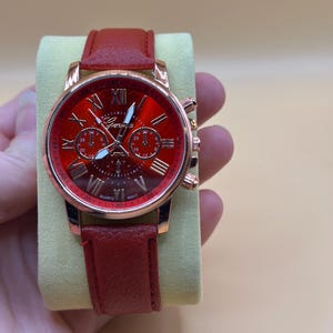 Könnte beinhalten: Eine rote Geneva-Quarzuhr mit rotem Lederarmband. Das Zifferblatt der Uhr ist rot mit goldenen römischen Ziffern und Zeigern. Die Uhr hat drei kleinere Zifferblätter und ein goldfarbenes Gehäuse.