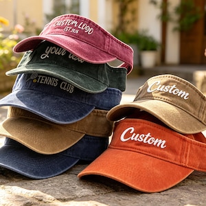 May include: A selection of visors in shades of burgundy, green, navy, tan, and orange. Each visor has a curved peak and features custom text in white. The visors are made of a soft, textured material.