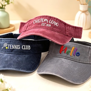 Könnte beinhalten: Drei Sonnenblenden in verschiedenen Farben: ein verwaschenes Rot mit "CUSTOM LOGO EST.2026", ein Marineblau mit "TENNIS CLUB" und ein graues mit "Hello" in Regenbogenfarben. Diese Visiere sind zum Schutz der Augen vor der Sonne konzipiert.