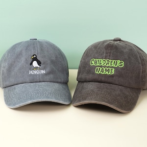 May include: Two baseball caps. One is grey with a penguin graphic and the word "PENGUIN" embroidered below. The other is dark grey with the words "CHILDREN'S NAME" in green embroidery. Both caps have a curved peak and adjustable strap.