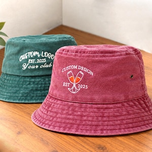 Peut inclure: Un bob rouge foncé avec une broderie blanche indiquant CUSTOM DESIGN EST 2025 et deux flûtes à champagne, placé devant un bob vert foncé avec une inscription blanche CUSTOM LOGO EST 2025 Your club brodée sur le devant du chapeau.