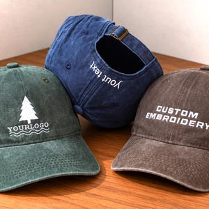 Könnte beinhalten: Drei Baseballkappen in Grün, Blau und Braun mit Used-Look. Die grüne Kappe zeigt ein weißes Tannenbaumsymbol und den Text "YOURLOGO". Die blaue Kappe hat "your text" auf der Rückseite eingestickt. Die braune Kappe trägt "CUSTOM EMBROIDERY".