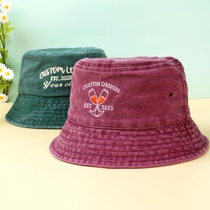 Könnte beinhalten: Zwei Eimerhüte, einer dunkelgrün und einer weinrot, werden gezeigt. Der grüne Hut hat einen weißen gestickten Text "CUSTOM LO EST. 2025 Your ol". Der weinrote Hut hat ein weißes Stickdesign mit zwei Sektgläsern und dem Text "CUSTOM DESIGN EST 2025".