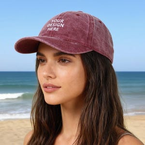Op de afbeelding: Een bordeauxrode baseballcap met de tekst "YOUR DESIGN HERE" in het wit. De pet wordt gedragen door een persoon op een strand met de oceaan en blauwe lucht op de achtergrond.