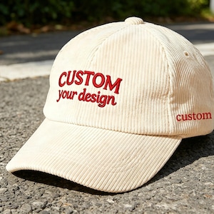Könnte beinhalten: Cremefarbene Cord-Baseballkappe mit den rot gestickten Worten "CUSTOM your design" auf der Vorderseite. Das Wort "custom" ist auf der Seite gestickt. Die Kappe hat einen gebogenen Schirm und einen Knopf oben.