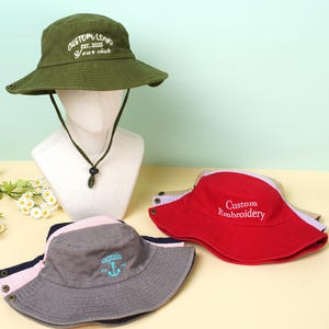 Peut inclure: Plusieurs chapeaux de pêcheur de couleurs variées, dont vert olive, rouge et gris. Le chapeau vert olive porte l'inscription "CUSTOM LOGO EST. 2025 Your club". Le chapeau gris affiche le mot "captain" et une ancre.