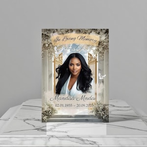 Peut inclure: Une photo commémorative avec le portrait d'une femme aux longs cheveux noirs, vêtue d'un haut blanc, avec le texte "In Loving Memory". La photo comprend le nom "Mandisa Mndai" et les dates 02.01.1955 ~ 20.09.2025, encadrée d'éléments floraux et architecturaux.