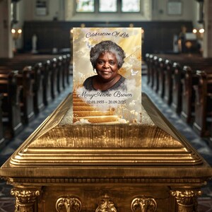 Könnte beinhalten: Ein Gedenkporträt mit dem Text "Celebration of Life" und "Mary Anne Brown 02.01.1955 ~ 20.09.2025" wird auf einem goldenen Sarg gezeigt. Das Porträt zeigt eine Frau mit grauem Haar vor dem Hintergrund von Wolken und Treppen.