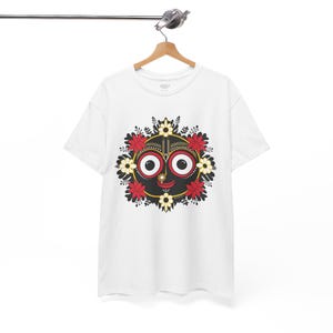 Può includere: T-shirt bianca con una grafica colorata di un volto con grandi occhi, un naso piccolo e una bocca sorridente. Il volto è circondato da fiori rossi e bianchi e foglie nere. La t-shirt è appesa a una gruccia di legno.
