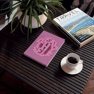 May include: A pink journal with the words "HEAL THY SELF" in a retro font. The journal is on a dark wooden table next to a cup of coffee and a travel magazine.