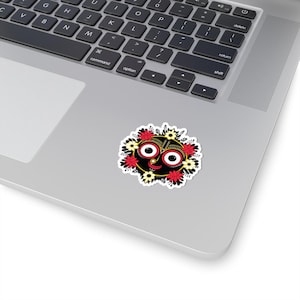 May include: A sticker featuring a stylized face with large, round eyes and a smiling red mouth. The face is black with gold accents and surrounded by red and yellow flowers. The sticker is on a laptop.