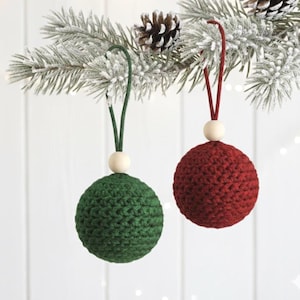 May include: Two handmade Christmas ornaments. One is a deep green, the other is a rich red. Each sphere is crocheted with a textured stitch, topped with a wooden bead and a loop for hanging. The ornaments are suspended from a snowy pine branch.