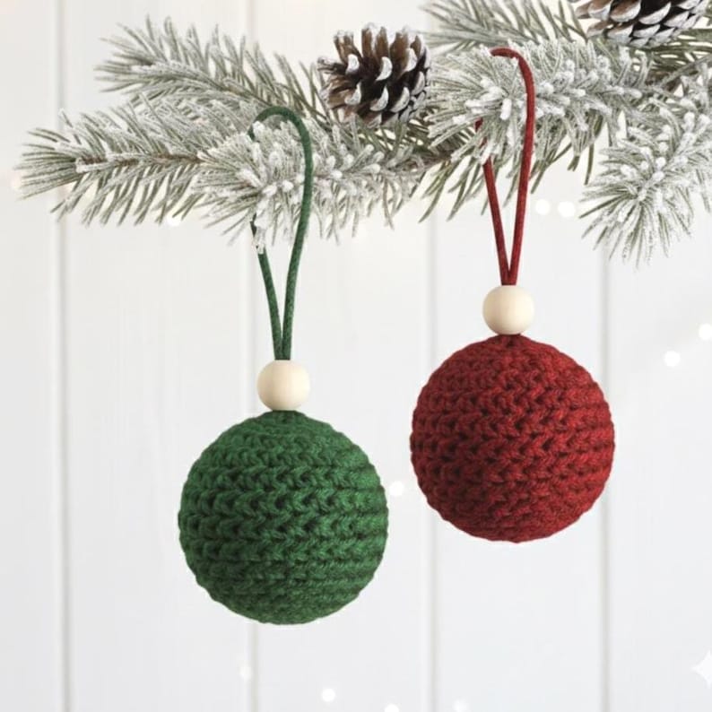 May include: Two handmade Christmas ornaments. One is a deep green, the other is a rich red. Each sphere is crocheted with a textured stitch, topped with a wooden bead and a loop for hanging. The ornaments are suspended from a snowy pine branch.