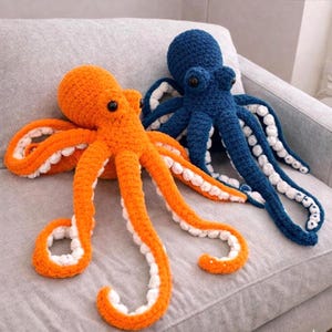 May include: Two crocheted octopus plush toys. One is bright orange with white accents on its tentacles, and the other is dark blue with white accents. Both have black eyes and are lying on a grey surface.