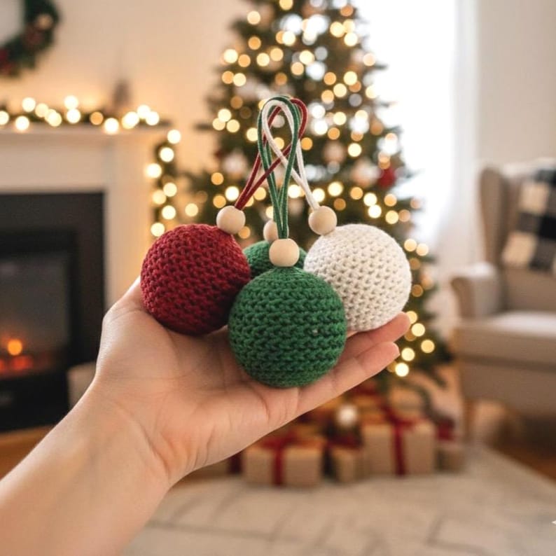May include: A hand holds three crocheted Christmas ornaments. The ornaments are red, green, and white, each with a wooden bead and a loop for hanging. A Christmas tree with twinkling lights is in the background.