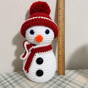 May include: A white crocheted snowman featuring a red hat with a pom-pom, a red and white scarf, blue eyes, and an orange nose. Two black buttons are on the body. A wooden ruler placed next to the snowman shows a height of 9 inches.