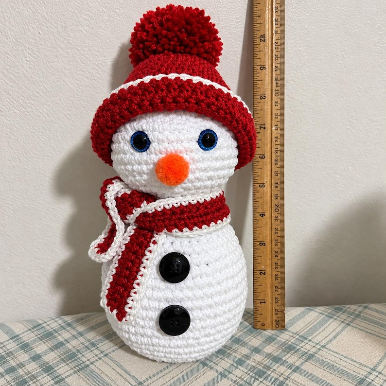 May include: A white crocheted snowman featuring a red hat with a pom-pom, a red and white scarf, blue eyes, and an orange nose. Two black buttons are on the body. A wooden ruler placed next to the snowman shows a height of 9 inches.