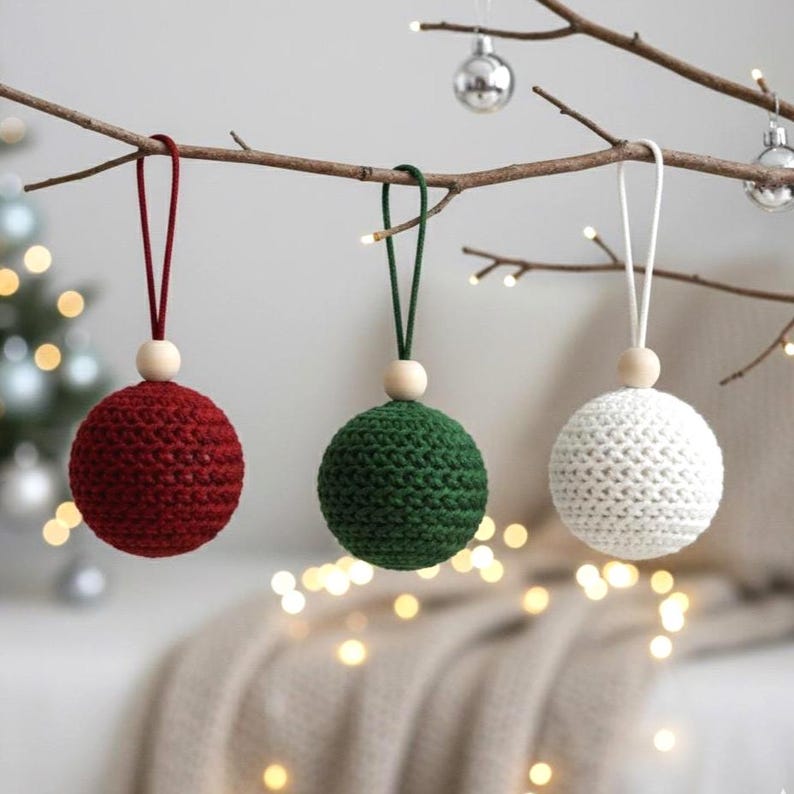 May include: Three handmade crochet Christmas ornaments in red, green, and white, each with a wooden bead and hanging loop. The ornaments are suspended from a tree branch, with soft lights and a blurred background.
