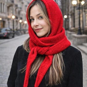 May include: A bright red knitted hood scarf featuring a textured stitch pattern that covers the head and wraps around the neck, tied in a knot at the front. The accessory is worn over a black coat, providing a vibrant contrast to the dark garment.