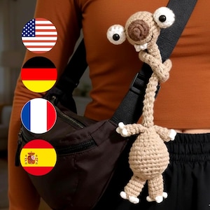 Könnte beinhalten: Eine handgefertigte Alien-Puppe aus Häkelarbeit in Beige und Weiß, mit großen weißen Augen und einem langen Hals. Die Puppe ist an einem schwarzen Riemen befestigt, mit einer braunen Tasche und einem orangefarbenen Oberteil im Hintergrund.