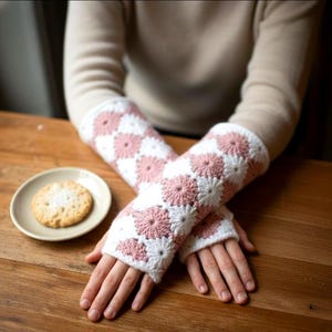 May include: Crocheted arm warmers in white and pink floral pattern. The arm warmers are made of a soft yarn and feature a daisy-like design. The cuffs are white. A small plate with a cookie is on a wooden table.