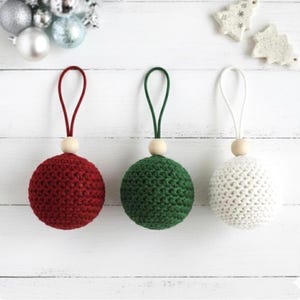 May include: Three handmade crochet Christmas ornaments in red, green, and white. Each sphere has a wooden bead and a loop for hanging. The ornaments are displayed on a white wooden surface.