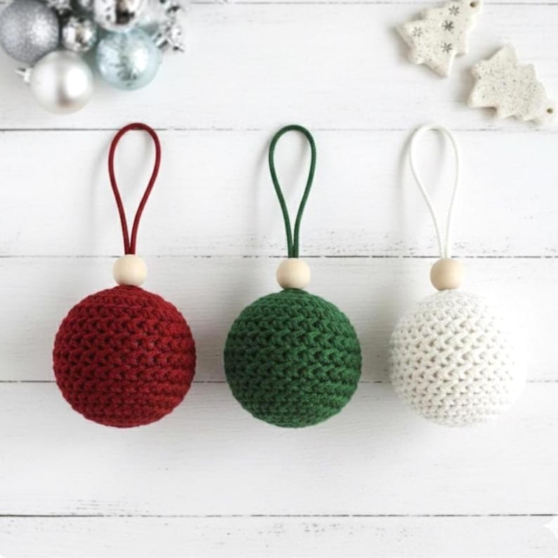 May include: Three handmade crochet Christmas ornaments in red, green, and white. Each sphere has a wooden bead and a loop for hanging. The ornaments are displayed on a white wooden surface.
