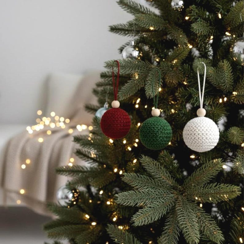 May include: Three handmade crochet Christmas ornaments in red, green, and white hang on a decorated Christmas tree. Each ornament is round with a wooden bead and a loop for hanging. The tree is adorned with lights and silver ornaments.