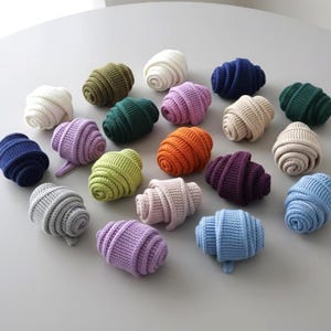 May include: A selection of colourful, knitted croissant-shaped objects in shades like white, green, blue, pink, orange, and purple. The objects are arranged on a light grey surface, highlighting their textured, ribbed design.