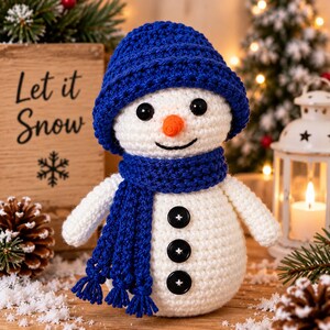 May include: A white crocheted snowman doll featuring a blue knit hat and a matching blue scarf with fringe. The snowman has black button eyes, a small orange carrot-shaped nose, a stitched black smile, and three black buttons on its torso.
