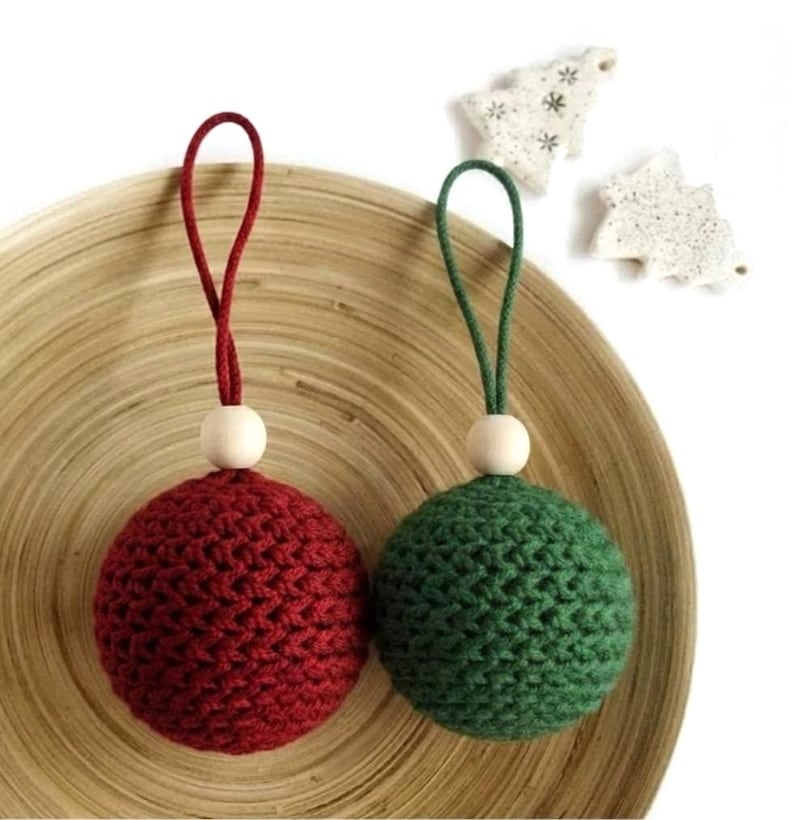 May include: Two crocheted Christmas ornaments. One is deep red with a matching cord and a wooden bead. The other is green with a green cord and a wooden bead. Two small white tree-shaped ornaments are in the background.