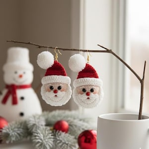 Peut inclure: Deux boucles d'oreilles faites à la main en crochet représentant le Père Noël, avec des bonnets rouges, des barbes blanches et des pompons, suspendues à une brindille. Les boucles d'oreilles ont des crochets dorés. Un bonhomme de neige et des décorations de Noël sont en arrière-plan.