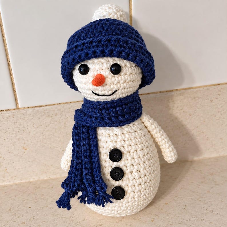 May include: A crocheted snowman doll made of off-white yarn, featuring a dark blue beanie with a pom-pom, a matching dark blue scarf with fringe, a small orange nose, black bead eyes, and three black buttons down the center of the body.