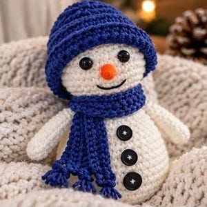 May include: A white crocheted snowman doll featuring a textured blue beanie hat and a matching blue scarf with fringe. The snowman has two black bead eyes, an orange conical nose, and three black buttons stitched vertically down its front.