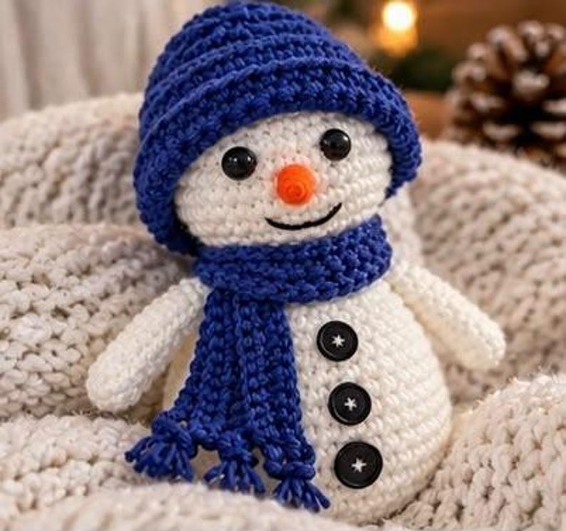 May include: A white crocheted snowman doll featuring a textured blue beanie hat and a matching blue scarf with fringe. The snowman has two black bead eyes, an orange conical nose, and three black buttons stitched vertically down its front.