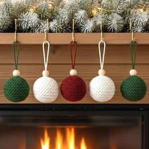 May include: Five handmade Christmas ornaments. Two are green, two are white, and one is red. Each ornament is a sphere made of knitted yarn, with a wooden bead and a hanging loop. The ornaments are hanging from gold hooks.