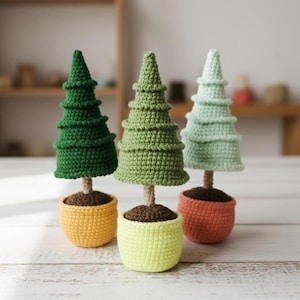 Peut inclure: Trois petits arbres de Noël crochetés. Chaque arbre est d'une nuance de vert différente, avec un tronc marron et un pot dans les tons jaune, vert et orange. Les arbres sont sur une surface blanche.