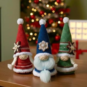 May include: Three handmade crochet gnomes in red, blue, and green. Each gnome has a conical hat with a white pom-pom, a cream-coloured face, and a white beard or trim. Decorative snowflakes adorn the hats and bodies.