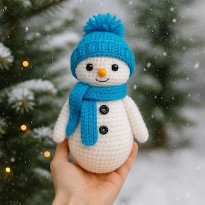 May include: A crocheted snowman figure, featuring a blue hat and scarf, black button details, and a carrot nose. The snowman is held in a hand, set against a snowy backdrop. The figure is handmade.
